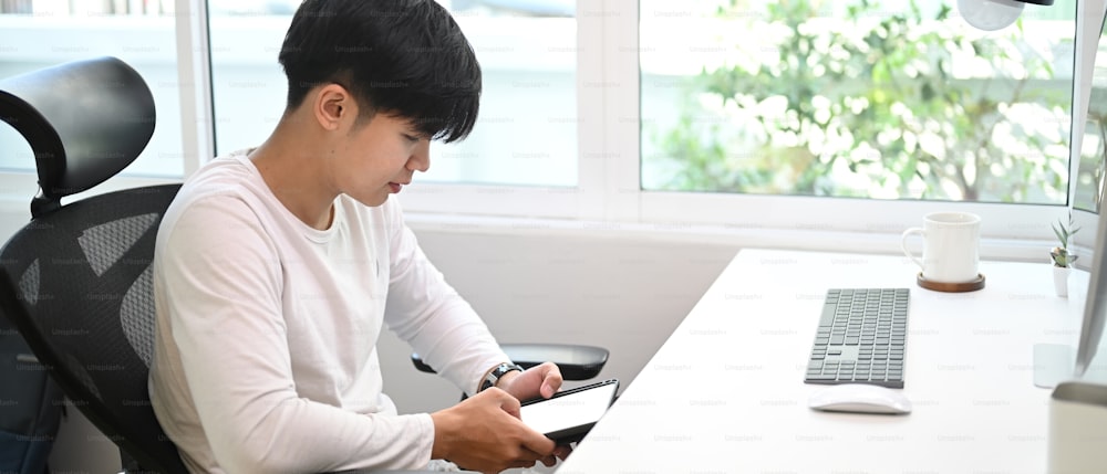 Imagen horizontal de un hombre independiente sentado frente a la computadora y usando un teléfono inteligente en la oficina en casa.