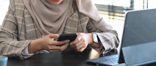 Foto cortada de empresária muçulmana de hijab usando smartphone e tablet enquanto estava sentada no escritório.