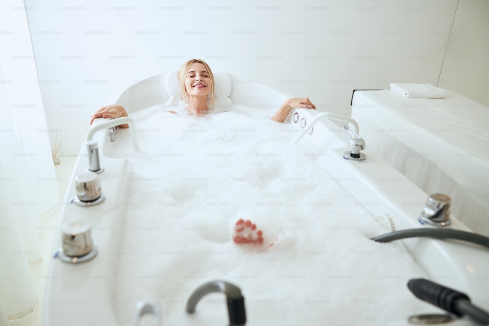 Retrato de corpo inteiro da senhora sorridente feliz relaxando no banho grande com espuma no salão de spa