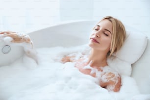 Portrait en gros plan d’une élégante femme caucasienne blonde fermant les yeux tout en se reposant dans le bain hydro avec de la mousse dans la station thermale