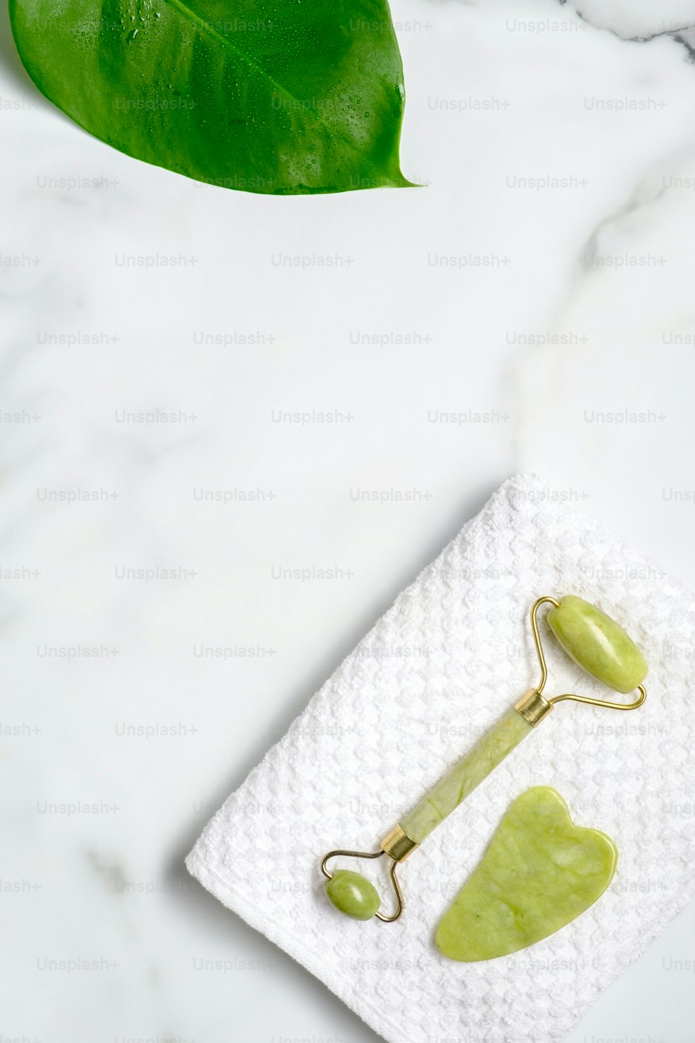 Jade stone facial roller and gua sha on towel on marble desk with green leaf. Facial skin care cosmetics set. Flat lay, top view.