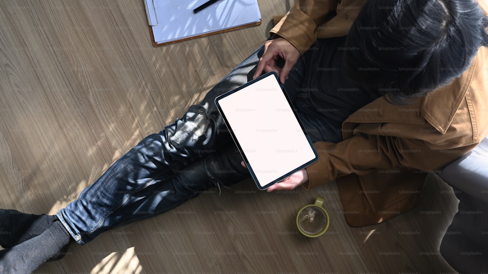 Over head shot of casual man freelancer sitting on floor and using digital tablet with blank screen.