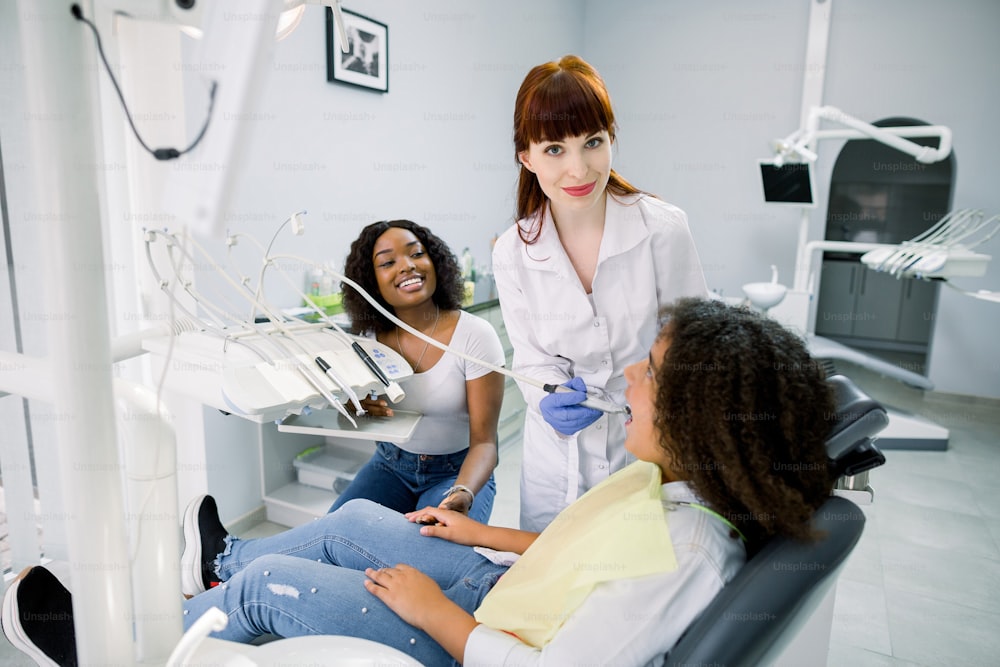 Señora africana madre y niña niña en cita en clínica moderna de odontología pediátrica. La doctora hace el tratamiento dental con taladro y espejo. Prevención de caries y restauración dental.