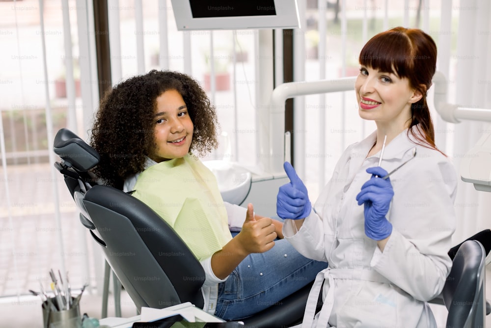 Pediatric dentistry, teeth care and orthodontics. Young Caucasian woman dentist, showing thumbs up together with her little patient, mixed raced teen girl, sitting in dental chair in modern clinic.
