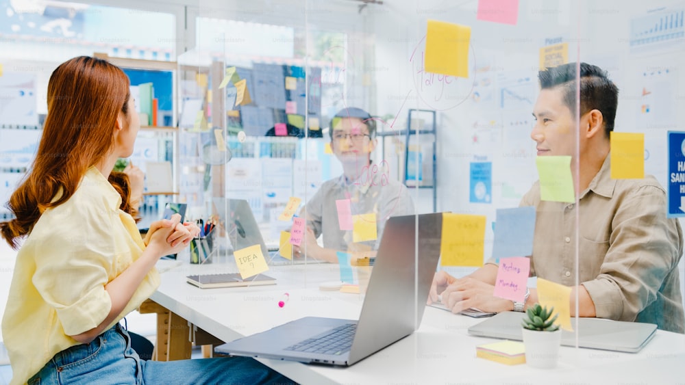 Asia businesspeople discussing business brainstorm meeting together share data and write on acrylic partition stand back in new normal office. Lifestyle social distancing and work after coronavirus.