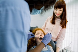 Back view of African female pediatric dental specialist making oral checkup of little curly red haired school girl, visiting dentist with her young mom. Dentist office, pediatric dentistry.