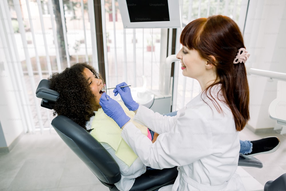 Dentist and kid at modern dental clinic. Young smiling caucasian dentist in white uniform and gloves, making examination of her small patient, little mixed raced curly girl, to prevent caries.