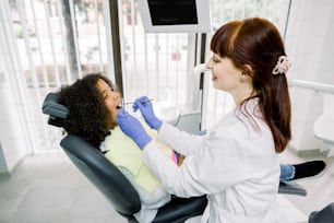 Dentista y niño en una clínica dental moderna. Joven dentista caucásica sonriente con uniforme blanco y guantes, examinando a su pequeña paciente, pequeña niña rizada de raza mixta, para prevenir la caries.