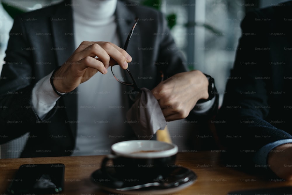 Abgeschnittenes Bild eines Mannes, der seine Brille putzt, während er Kaffee trinkt, während er sich auf ein Meeting vorbereitet