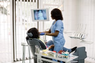 デンタルケア。小さな患者の歯の検査をするかわいいアフリカの女性医師、歯科の椅子に座っているアフリカ系アメリカ人の女の子、そして彼女の歯のデジタル画面に画像を表示しています