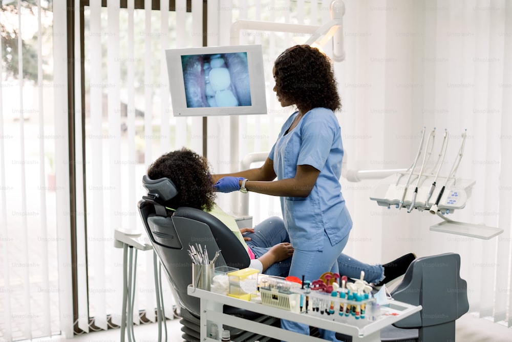 Dental care. Pretty African female doctor making examination of teeth of little patient, african american girl sitting in dentistry chair, and showing on the digital screen image of her teeth