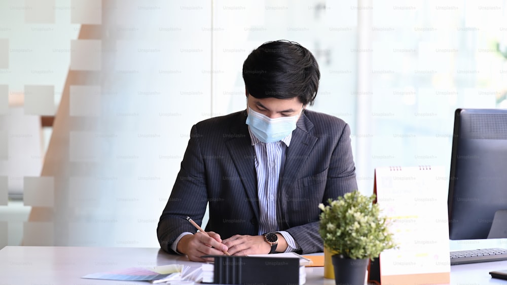 Young businessman wearing face mask concentrate writing information on document while sitting in office room.