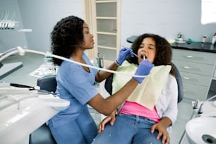 Foto de jovem e bonita dentista africana em uniforme azul, fazendo check-up dentário e tratamento de cárie com broca para seu pequeno paciente bonito, menina encaracolada de corrida mista, sentada em cadeira odontológica.