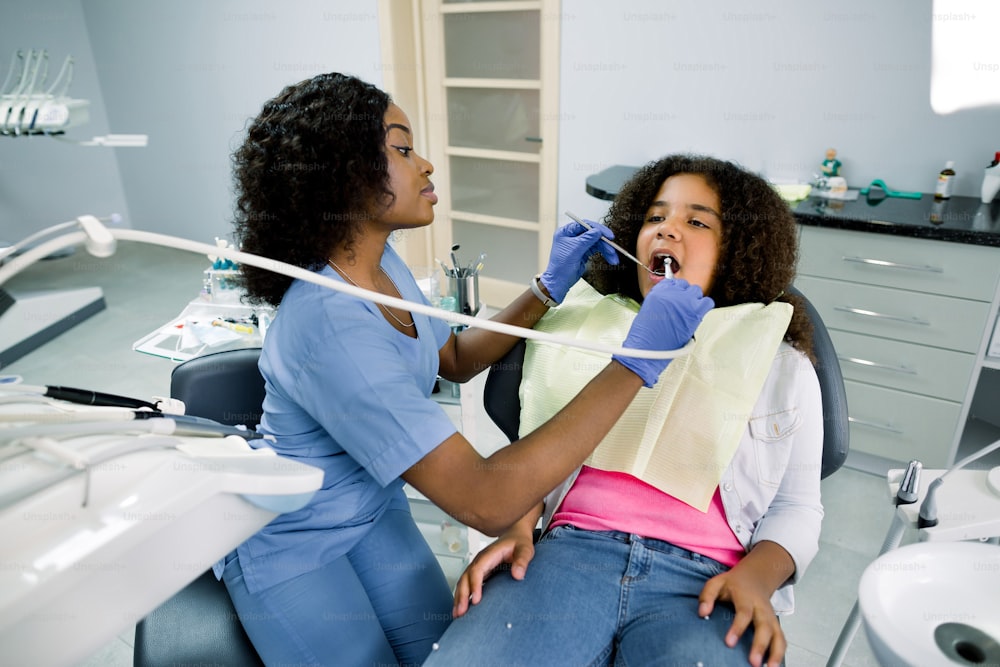 Foto de la joven y bonita dentista africana en uniforme azul, haciendo el chequeo dental y el tratamiento de la caries con taladro para su pequeña paciente linda, niña rizada de raza mixta, sentada en el sillón dental.