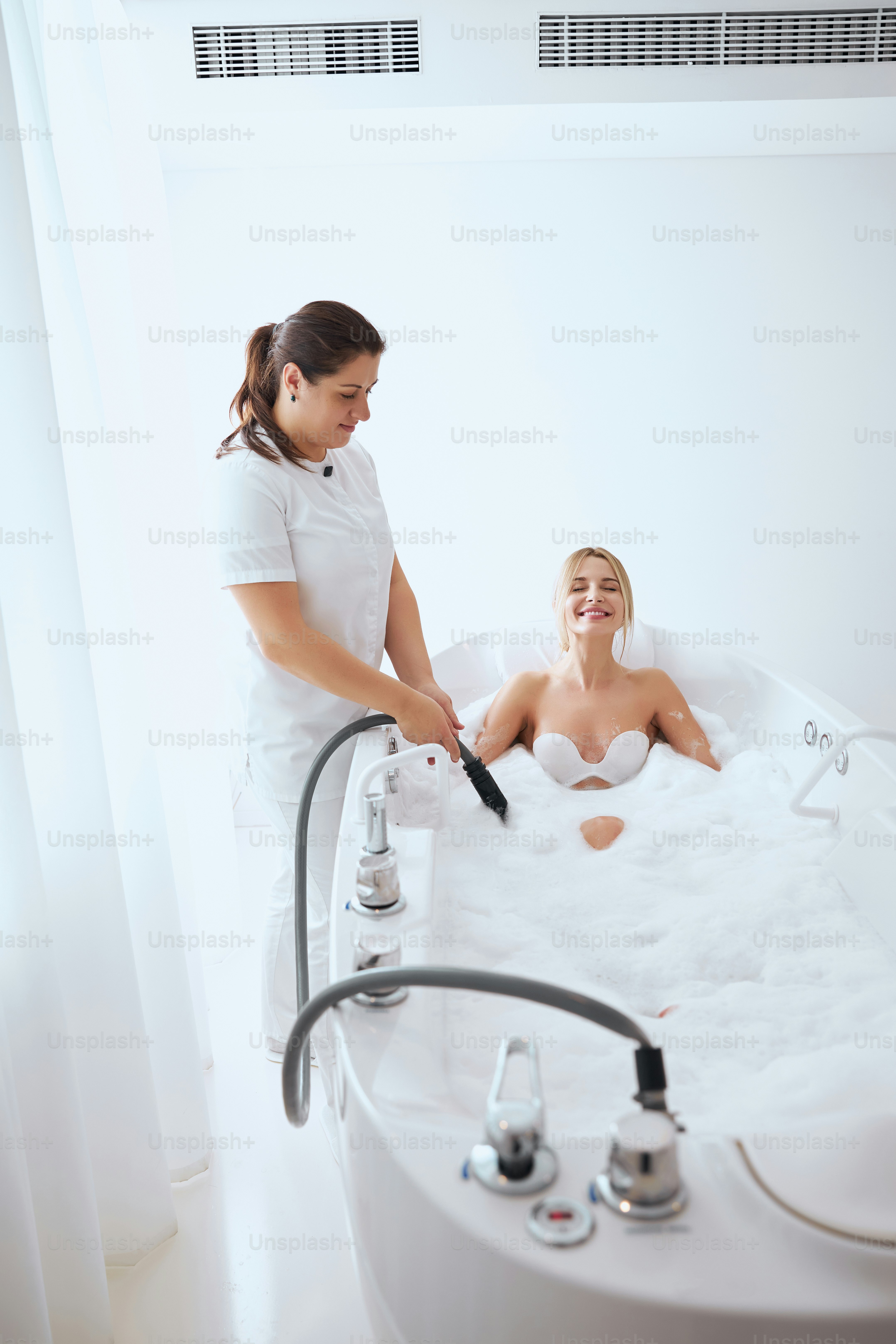 Side view portrait of charming female beautician using device for underwater massage while young lady lying in bathtub