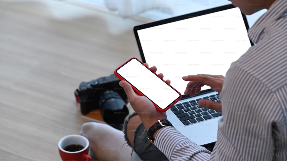 Close up view of man photographer using mock up laptop and smart phone with blank screen while sitting on wooden floor.