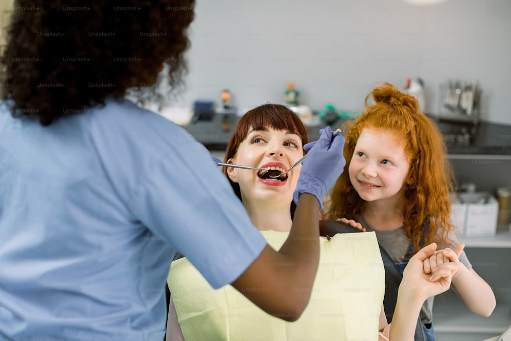 Close up of young lady, sitting in dental chair, supporting by her little daughter, while black female dentist cures her teeth. Back view of dentist wearing latex gloves and holding tools in hands.