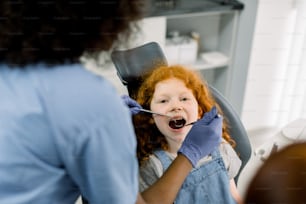 Vista superior de cerca de una bonita niña pelirroja rizada en la silla del dentista, con la boca abierta, teniendo un examen dental. Vista posterior de las manos de una dentista africana con guantes, sosteniendo herramientas.