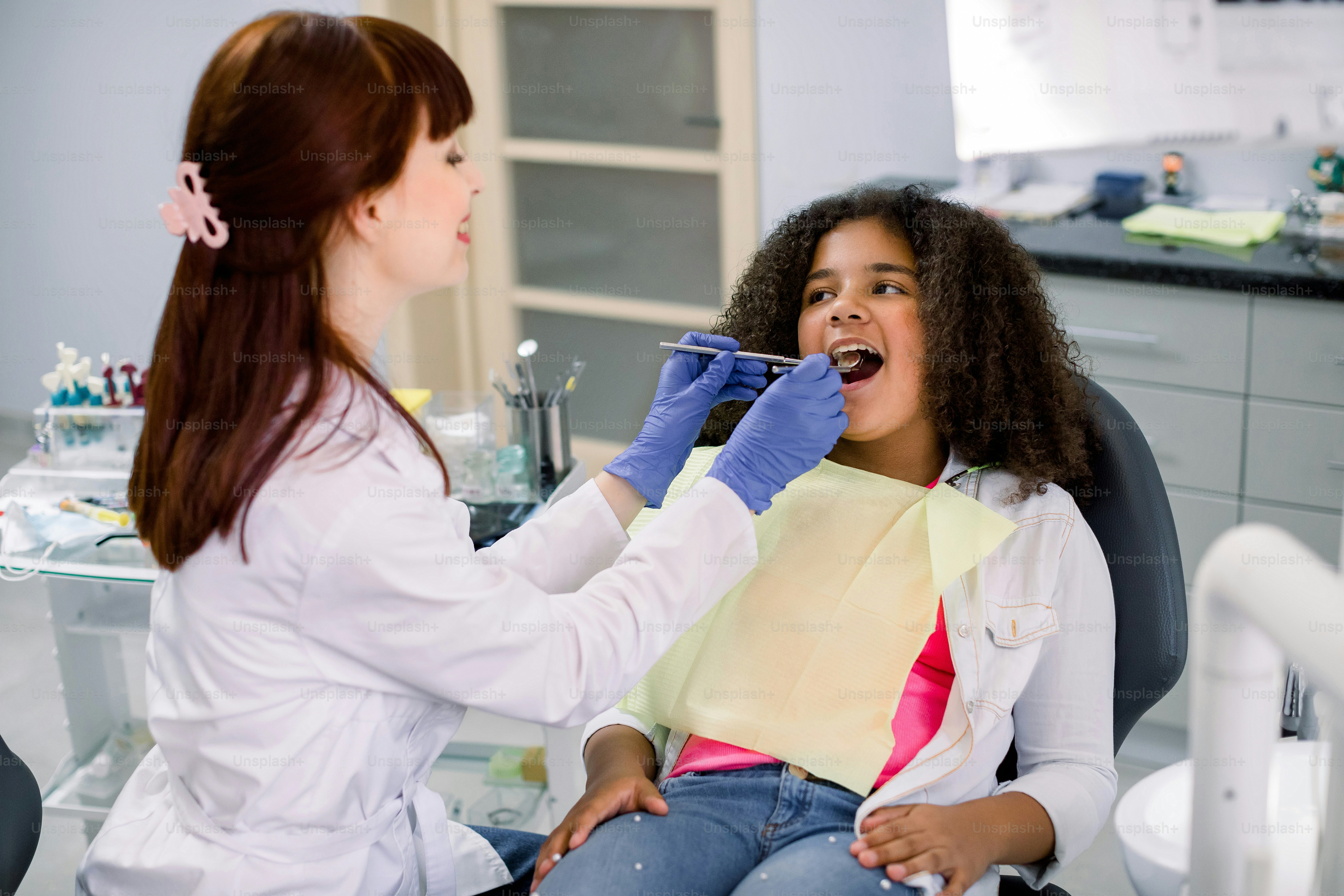 Ziemlich ein gemischtrassiges Teenager-Mädchen im Zahnarztstuhl, mit offenem Mund, zahnärztlich untersucht. Seitenansicht einer lächelnden kaukasischen Zahnärztin in Handschuhen, die zahnärztliche Werkzeuge hält.