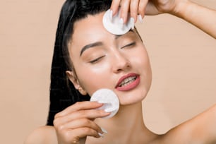 Young asian woman with eyes closed, enjoying cleaning of face, using cotton pads with cleansing lotion or facial toner for removing makeup. Skin care concept. Beige background, copy space.