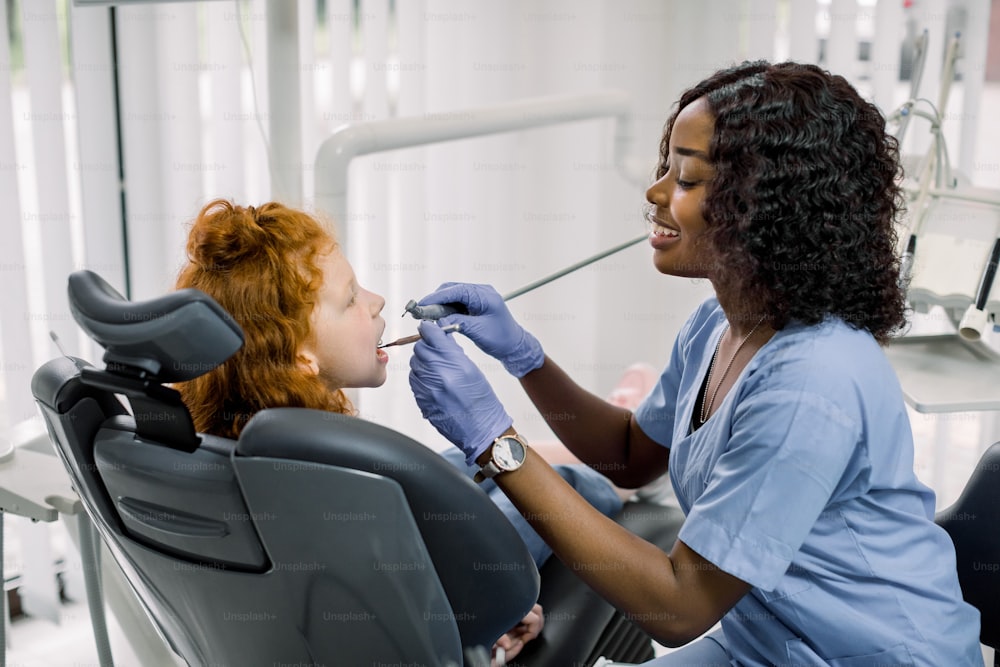 Side view of happy pretty african lady dentist in blue uniform and gloves, making examination or curing caries and toothache for her little patient, red haired curly girl, sitting in dental chair.