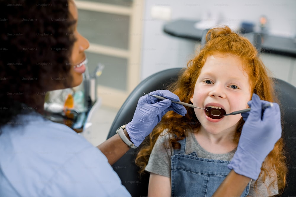 Un niño en una clínica dental. Retrato de cerca de una niña pequeña con el pelo rojo rizado, sentada con la boca abierta en el sillón dental mientras que el dentista africano femenino hace el chequeo de los dientes usando el espejo dental