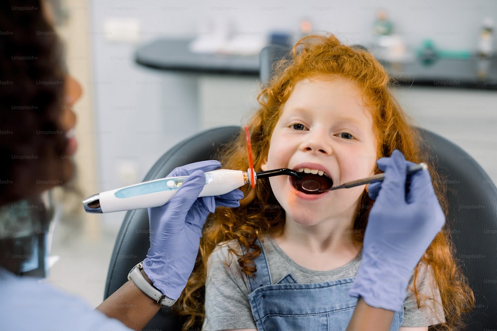 Close up view of little red haired girl patient having dental treatment at dentist's office. Young female african dentist making tooth restoration with filling using curing polymerization UV lamp.