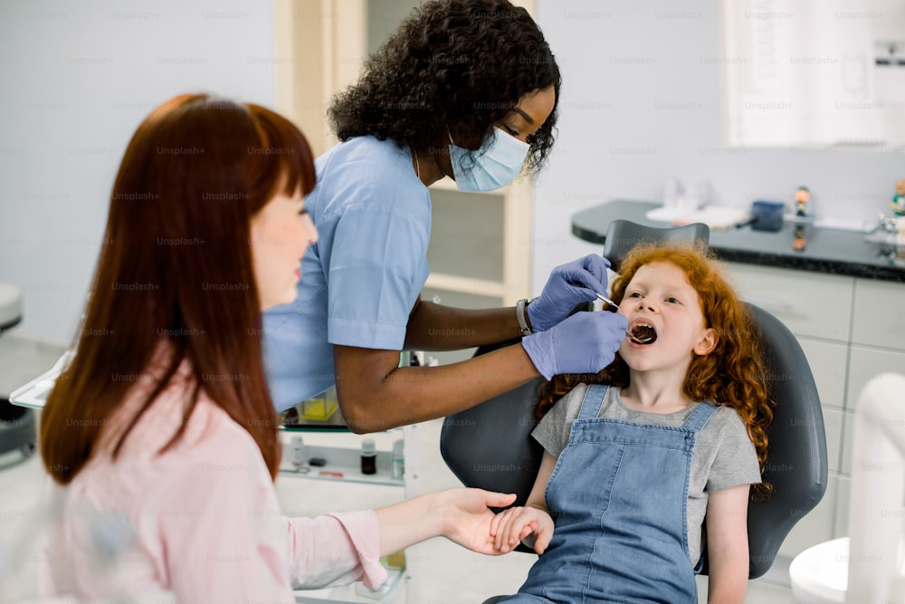 Cute schoolgirl seating in dentist chair and holding mom's hand, while professional African woman dentist providing regular teeth checkup. Caries prevention and teeth hygiene.