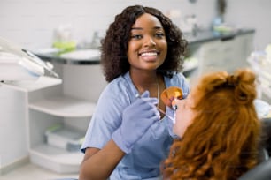 Smiling black woman dentist wearing blue uniform, providing tooth restoration and filling with curing polymerization UV lamp for her little patient, cute girl with red curly hair.