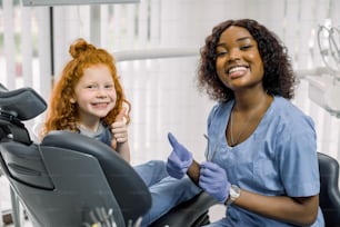 Front view of cheerful smiling African female dentist looking at camera, and little red haired girl patient, sitting in dentist chair, showing thumbs up. Concept of teeth health, oral care.