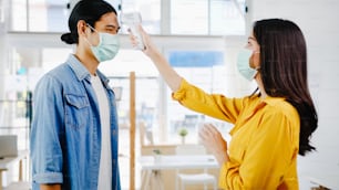 Asia female receptionist conducting wear protective face mask use infrared thermometer checker or temperature gun on customer's forehead before enter office. Lifestyle new normal after corona virus.