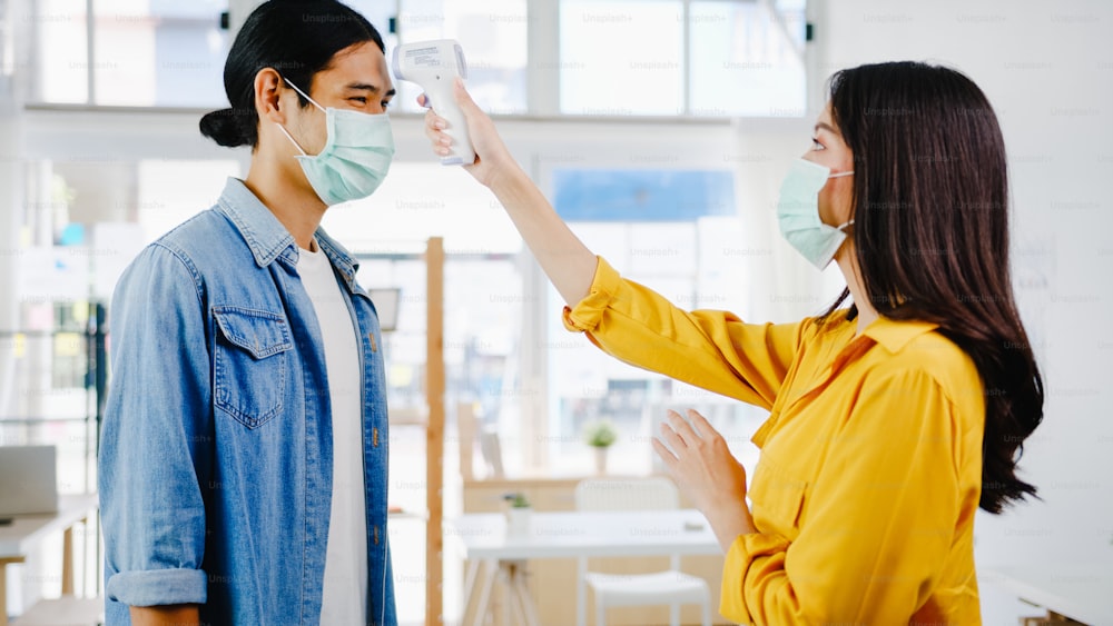 Asien weibliche Rezeptionistin trägt Schutzmaske Gesichtsmaske verwenden Infrarot-Thermometer-Checker oder Temperaturpistole auf der Stirn des Kunden vor dem Betreten des Büros. Lebensstil neue Normalität nach Coronavirus.