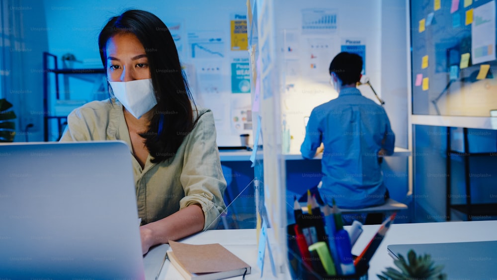 オフィスの夜に仕事に戻ってラップトップを使用しながら、ウイルス予防のための新しい通常の状況で社会的距離のために医療用フェイスマスクを着用した幸せなアジアのビジネスマン。コロナウイルス後の生活と仕事。