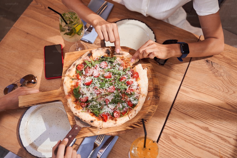 Summer dinner or lunch. Flat-lay of female hands taking freshly baked Italian pizza with vegetables and fresh basil over wooden table. Top view