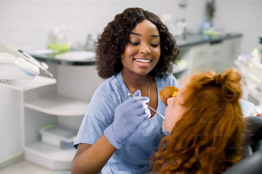 Pretty african female dentist in blue uniform and gloves, making tooth restoration and filling with curing polymerization UV lamp for her little patient, cute girl with red curly hair.