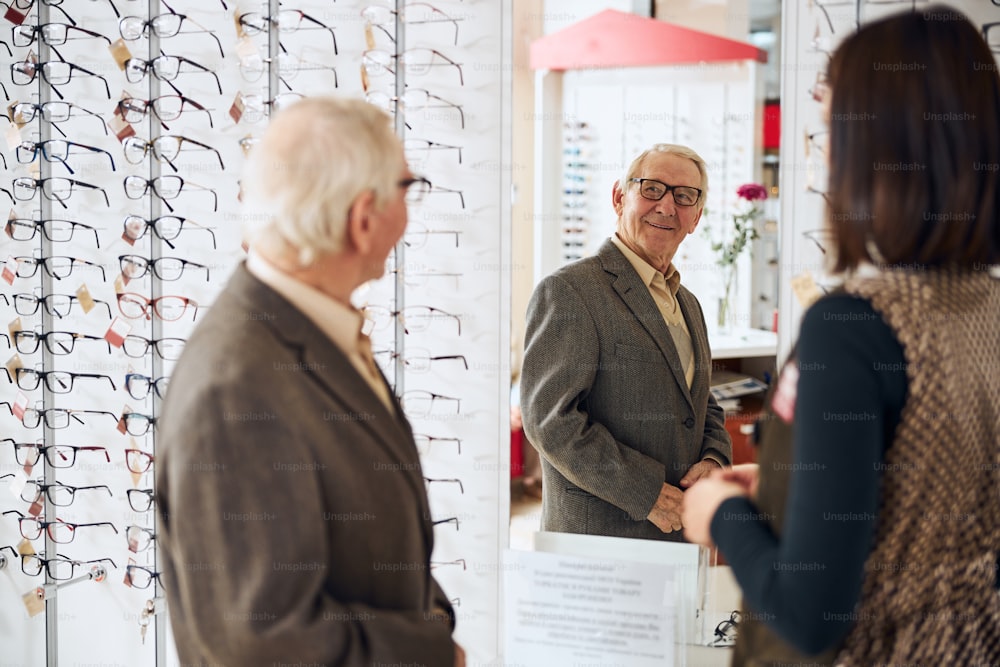 Attractive pleased elderly male customer in trendy prescription eyeglasses looking at himself in the mirror