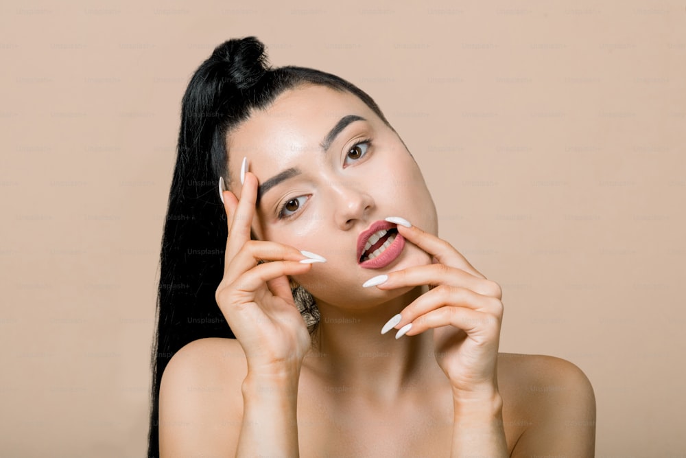 Beautiful young sexy Asian woman with ponytail hairstyle, posing to camera with hands touching face, lips and eyebrows, isolated on beige background. Close up portrait. Beauty and skin care concepts.