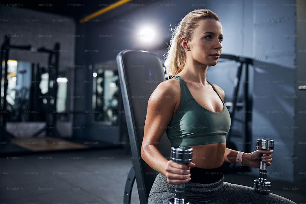 Decisive sportswoman getting ready for exercises while clenching on the dumbbells on a fitness equipment