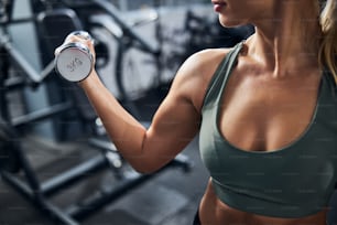Lady practicing bodybuilding among the gym machines while raising one metal dumbbell with a muscular hand