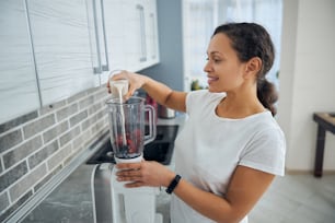 Smiling young female adding the protein-rich ingredient to the blender container filled with the fruit