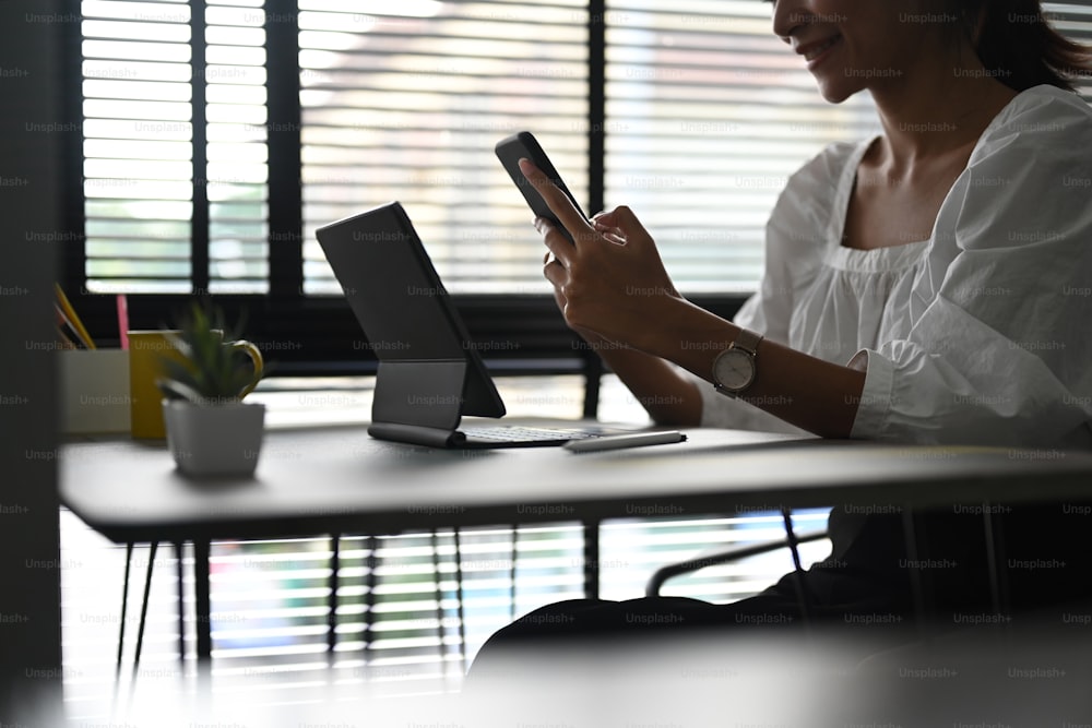 Colpo ritagliato di giovane femmina seduta davanti al suo tablet computer in ufficio moderno e utilizzando il telefono cellulare.