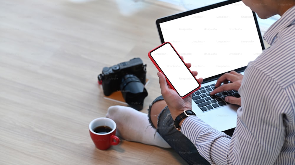 写真家やフリーランスが、木製の床に座りながら、画面が空白のモックアップラップトップとスマートフォンを使用しています。