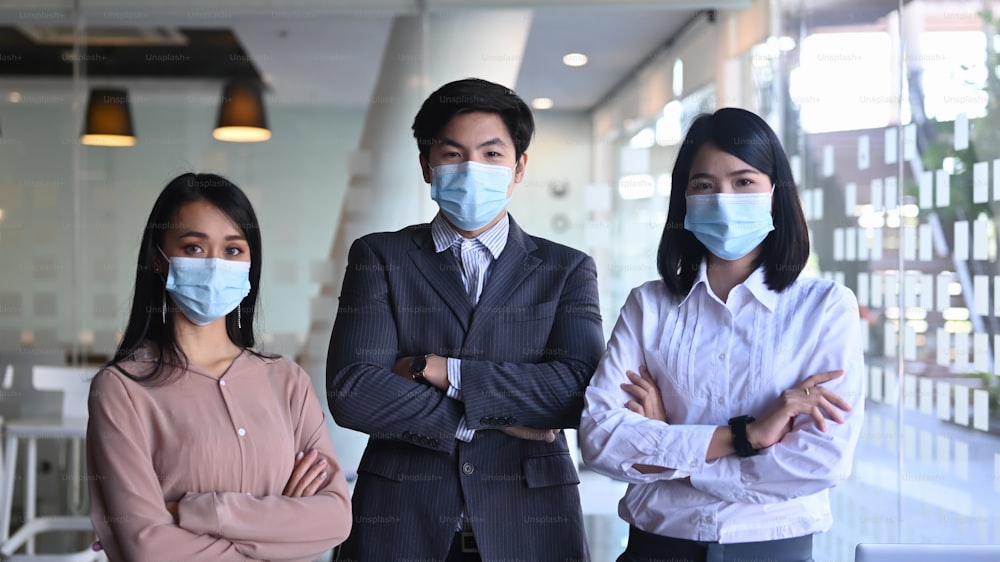 현대 사무실에서 팔짱을 끼고 서 있는 동안 의료용 마스크를 쓴 자신감 있는 사업가들.