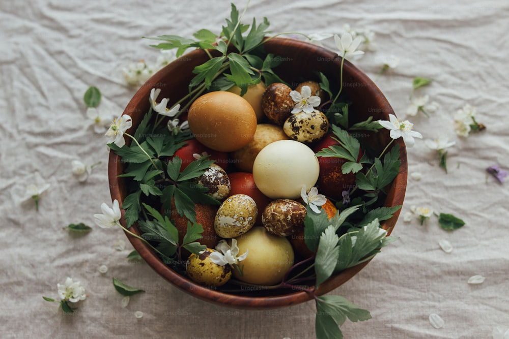 イースターおめでとう!素朴なリネンの背景に木製のボウルに春の花とイースターエッグ。審美的な季節のグリーティングカード。天然染料と春の花のスタイリッシュなイースターとウズラの卵。