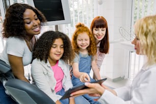Kinder und Mütter in der Zahnarztpraxis. Zwei multiethnische Mütter mit ihren jugendlichen Töchtern, die auf einem Zahnarztstuhl sitzen und auf einen digitalen Tablet-PC in den Händen einer blonden Zahnärztin zeigen.