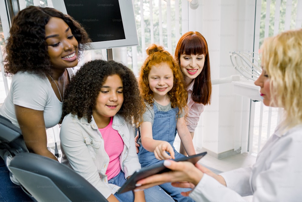 Children and mothers at dentist's office. Two multiethnic mothers with their teen daughters, sitting in dentistry chair, pointing on digital tablet pc in hands of female blond dentist.