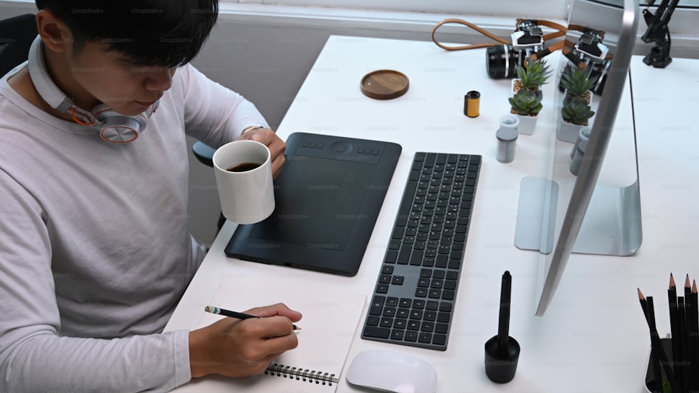 High angle view of young man photographer drinking coffee and making note at his workstation.