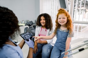 Children at dentist's office. Two multiethnic teen girls, sitting in dentistry chair, looking at camera and pointing on tablet with teeth panoramic scan together with her female african dentist.