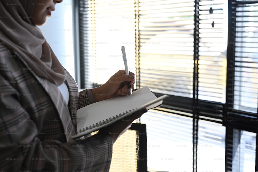 Muslim businesswoman in hijab hand holding a pen writing on notebook while standing in front of window.