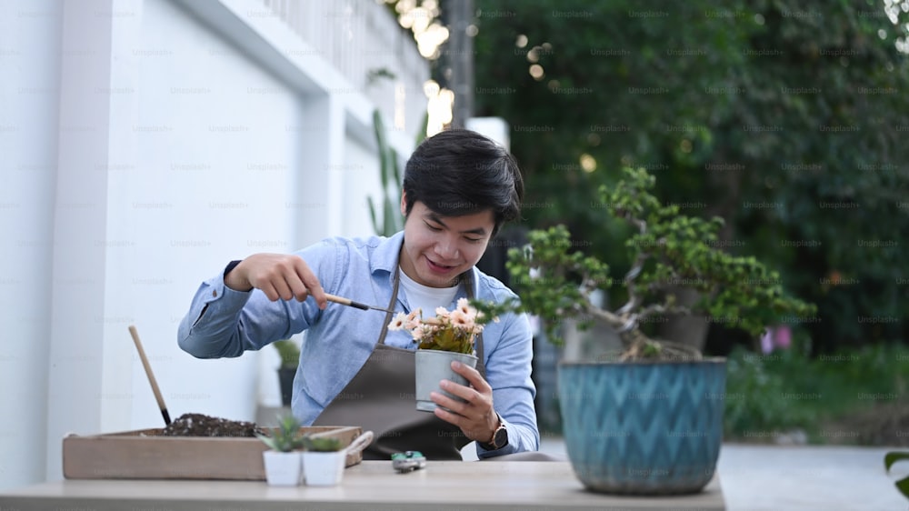 A cheerful young man planting flowers in pot with dirt or soil at home.